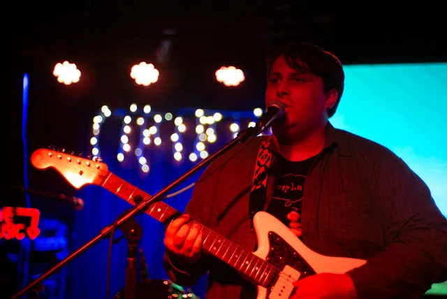Luke Evans performing live with a white electric guitar under red and cyan stage lights.